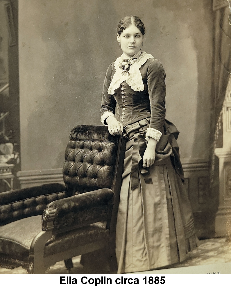 Ella Coplin circa 1885; black and white studio photo of a young woman in an ornate outfit consisting of a velour bodice with large white lace collar and silk flowers at the neck and a long satin skirt with multiple ruffles, dark curled hair parted in the middle and pulled back into a bun, a slight smile on her closed lips as she stares directly into the camera. She stands behind a velour-upholstered armchair, her right forearm resting on its back.